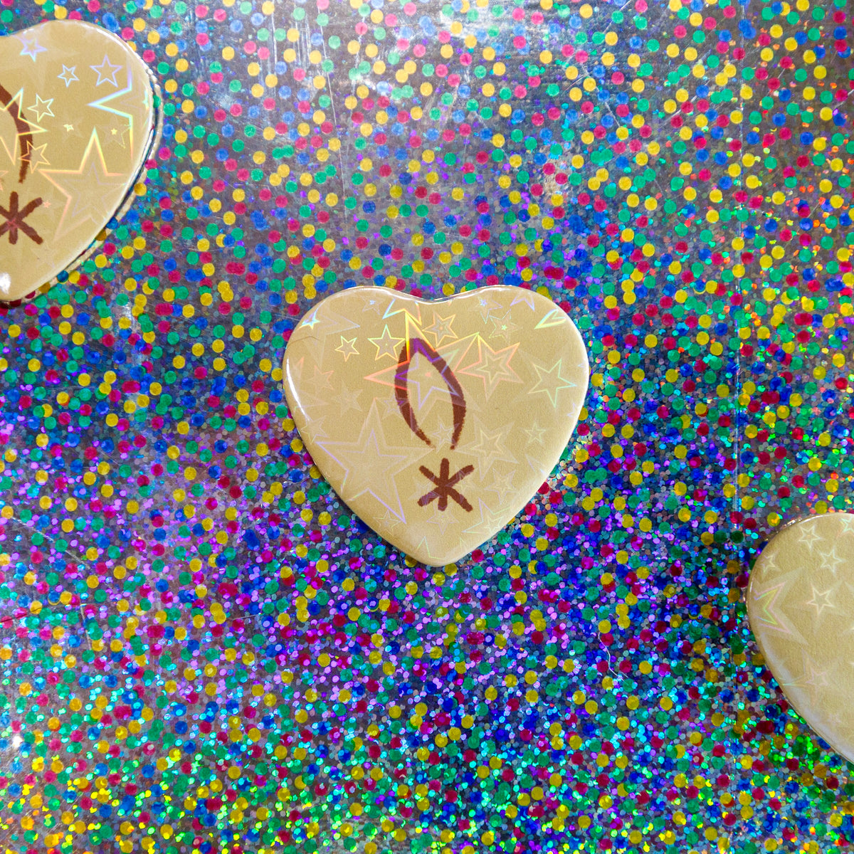 A heart-shaped beige pin with a brown crescent and star symbol, displayed on a vibrant, multicolored holographic glitter background.